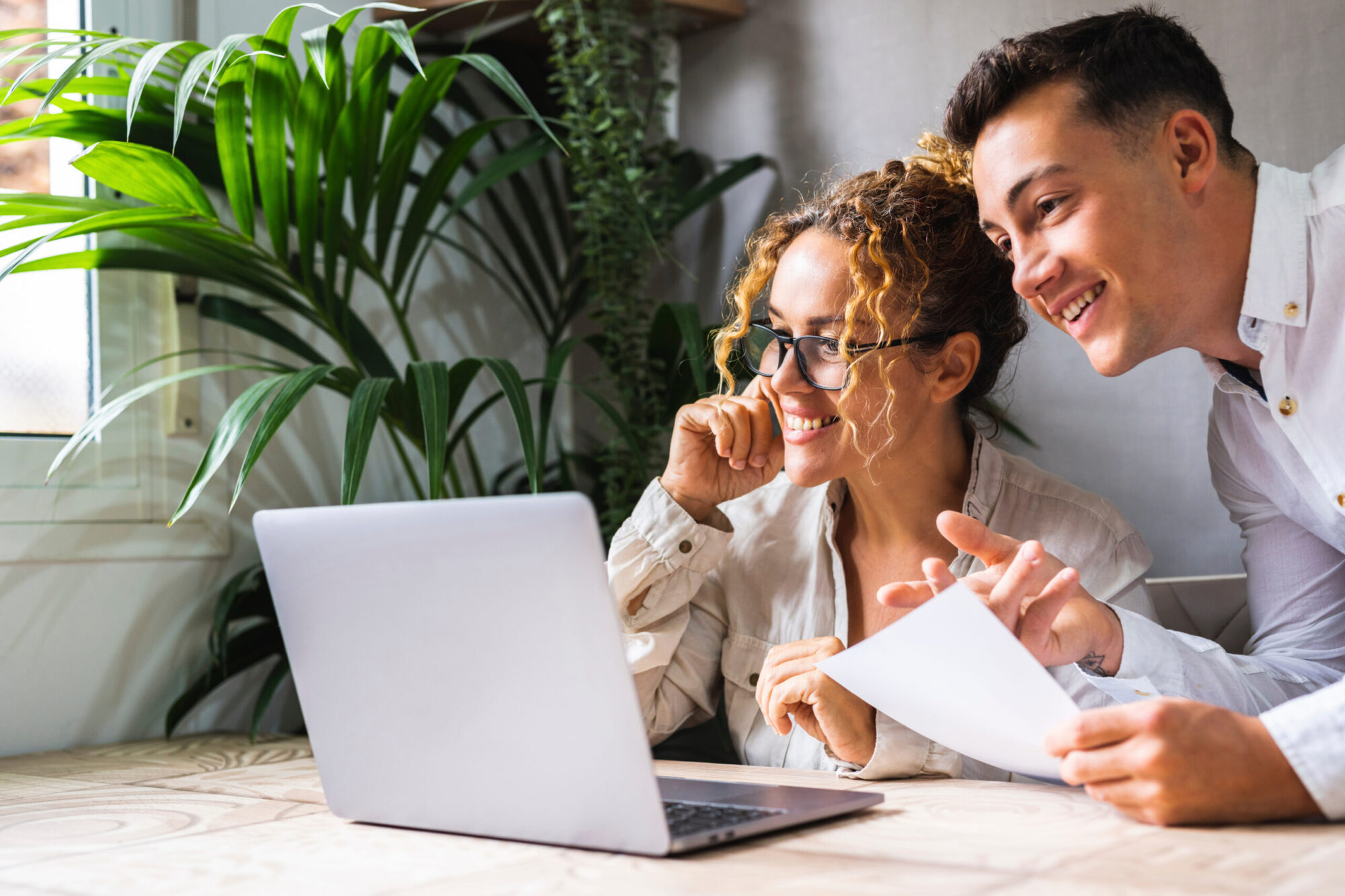 Two people working together with computer at the desk in home office workplace. One young man and adult woman smile and enjoy small business. Entrepreneur with assistant using computer wireless online